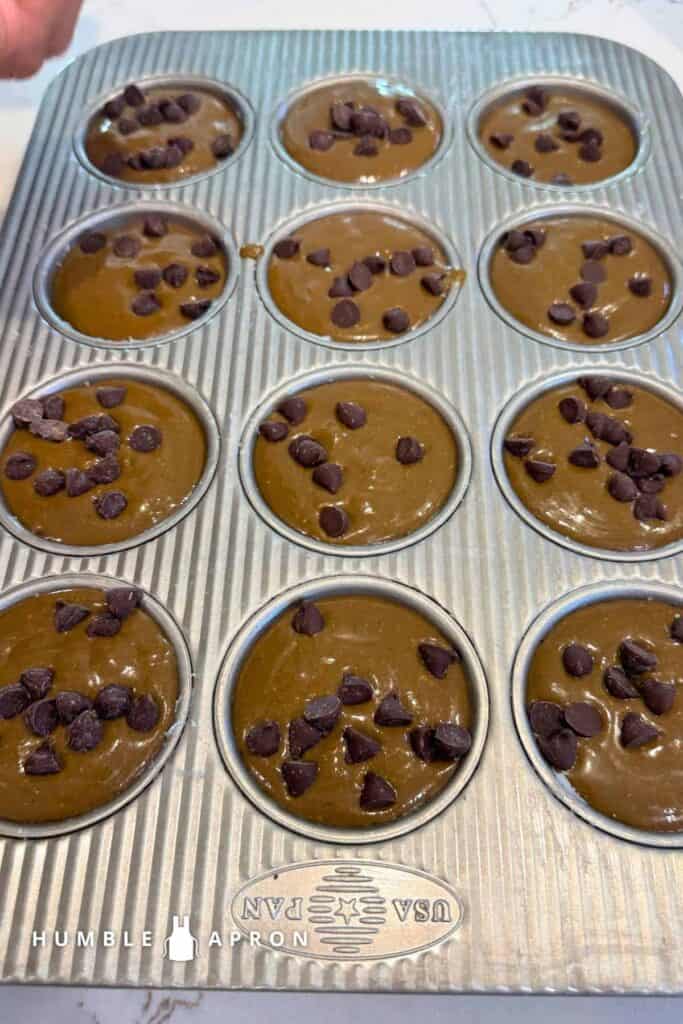 A metal muffin tin filled with chocolate chip muffin batter, ready to bake. Each cup is topped with chocolate chips. The words HUMBLE APRON and a small logo are in the lower left corner.