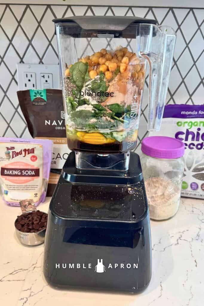 A blender filled with chickpeas, spinach, eggs, Greek yogurt, and other ingredients for chocolate chickpea muffins, surrounded by cocoa powder, chia seeds, protein powder, and chocolate chips.