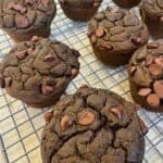 Chocolate muffins with chocolate chips on top are cooling on a wire rack on a kitchen counter. The muffins look moist and rich, and there is a Humble Apron watermark in the lower left corner.
